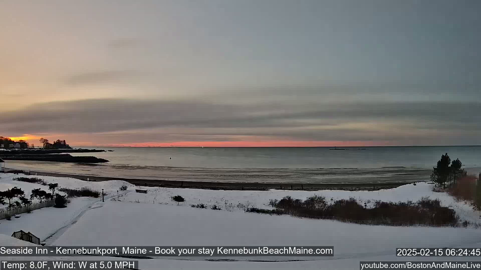 A snow-covered beach and coastline are visible at sunrise, with a calm ocean and a pinkish-orange sky.
