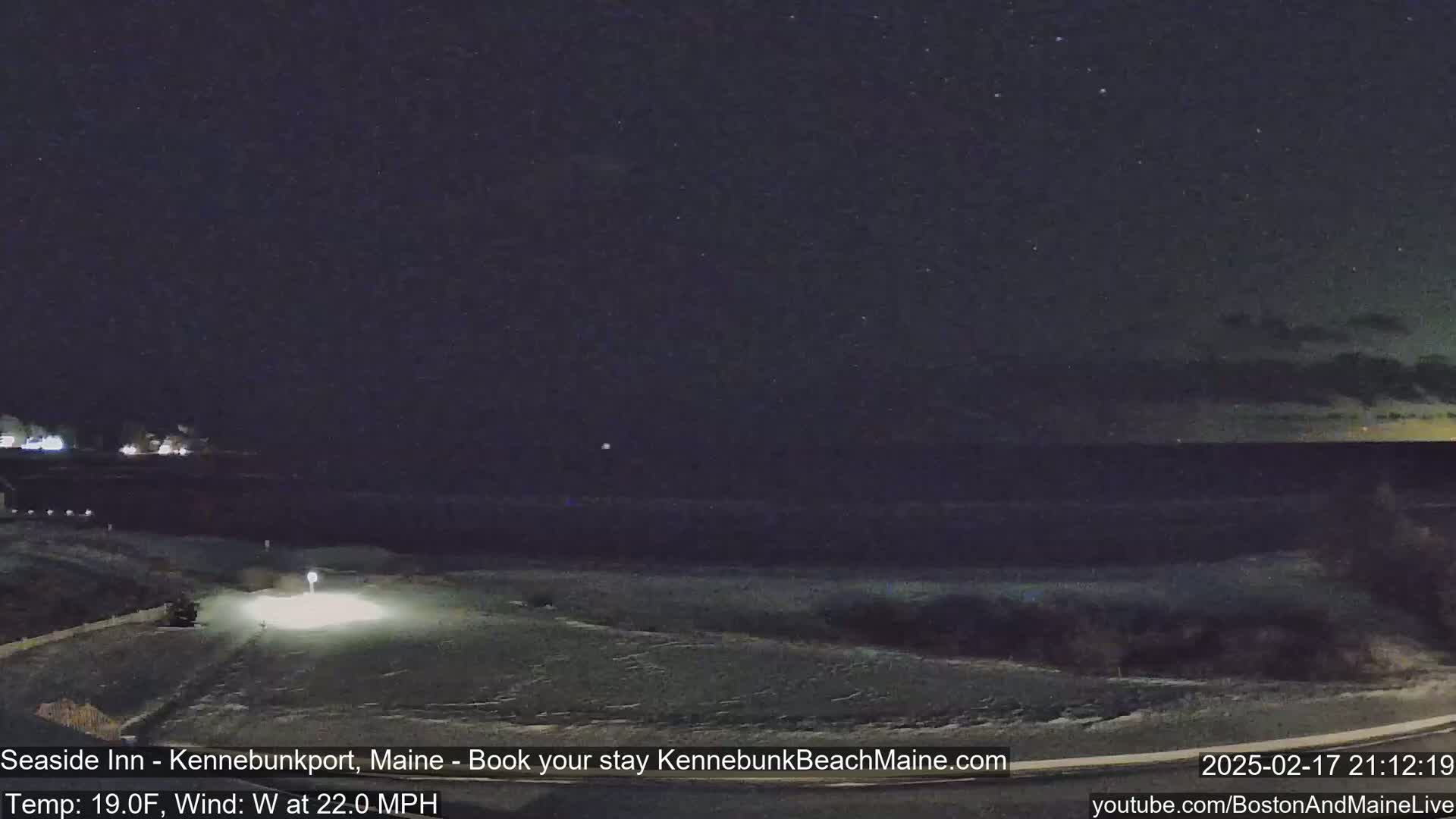 A nighttime coastal scene shows a dark, starry sky, a snow-covered shoreline, and a vehicle with headlights on driving along a road near the water under windy, nineteen-degree Fahrenheit conditions.