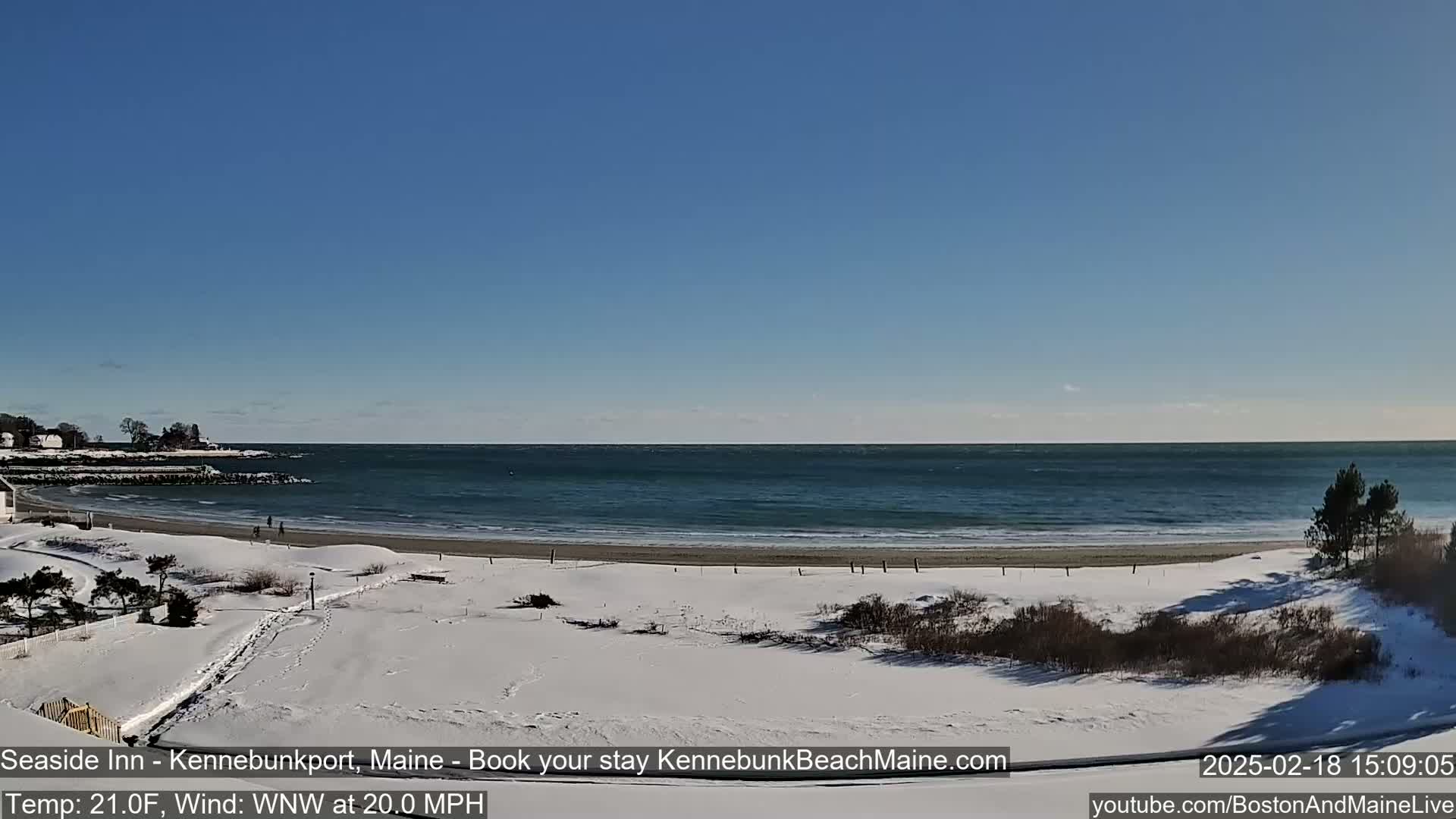 A snow-covered beach with a partly cloudy blue sky and a wavy ocean under a windy, 21°F temperature.