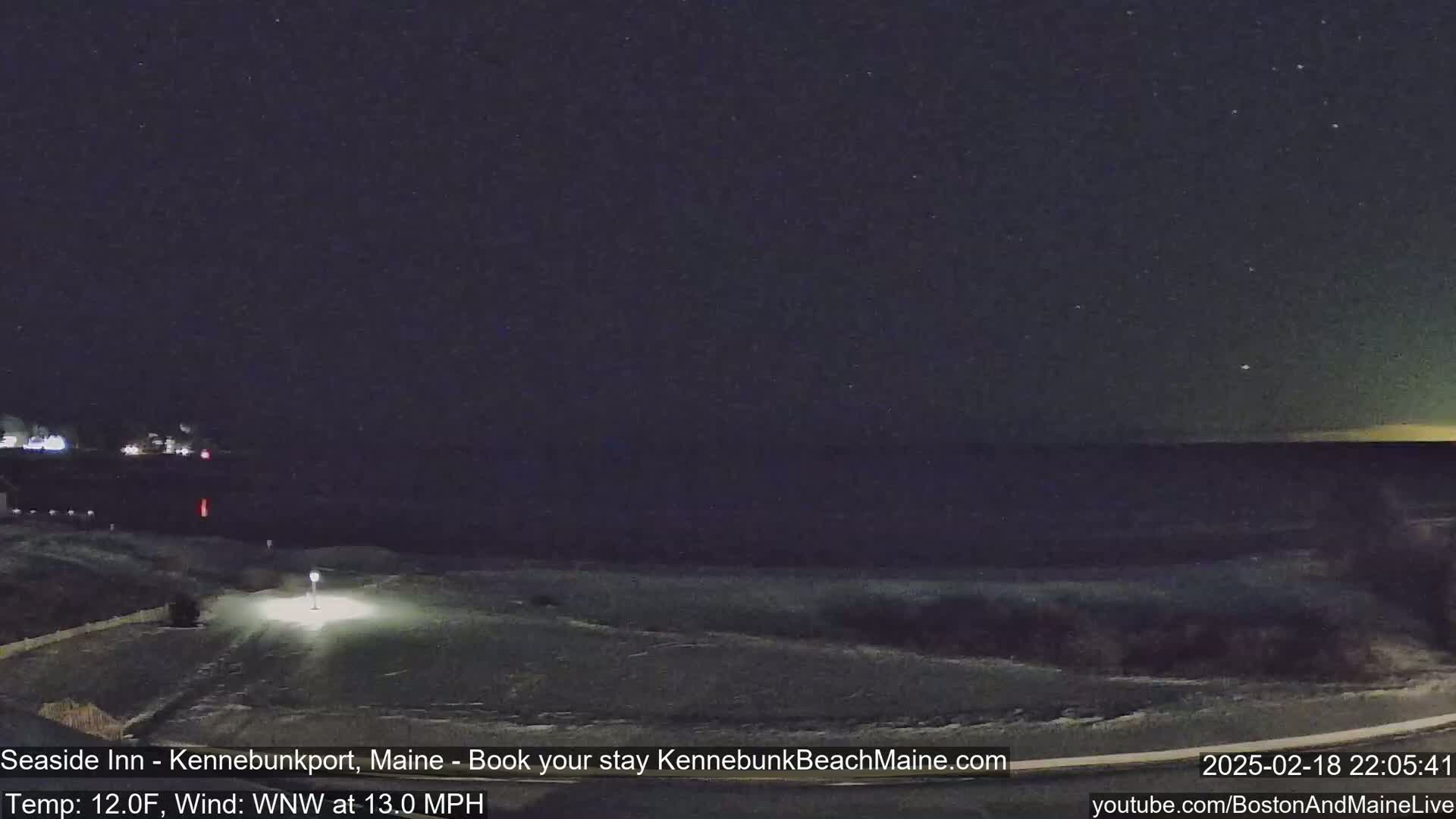 A nighttime, snow-covered coastal scene shows a dark ocean under a starry sky, with a road and faint lights visible in the foreground.