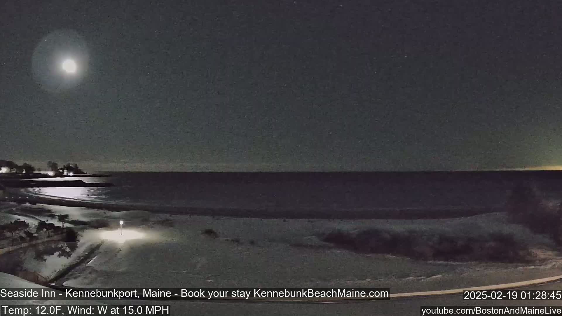 A nighttime view of a snow-covered beach with a calm ocean under a bright moon, with a 12°F temperature and a 15 mph west wind.