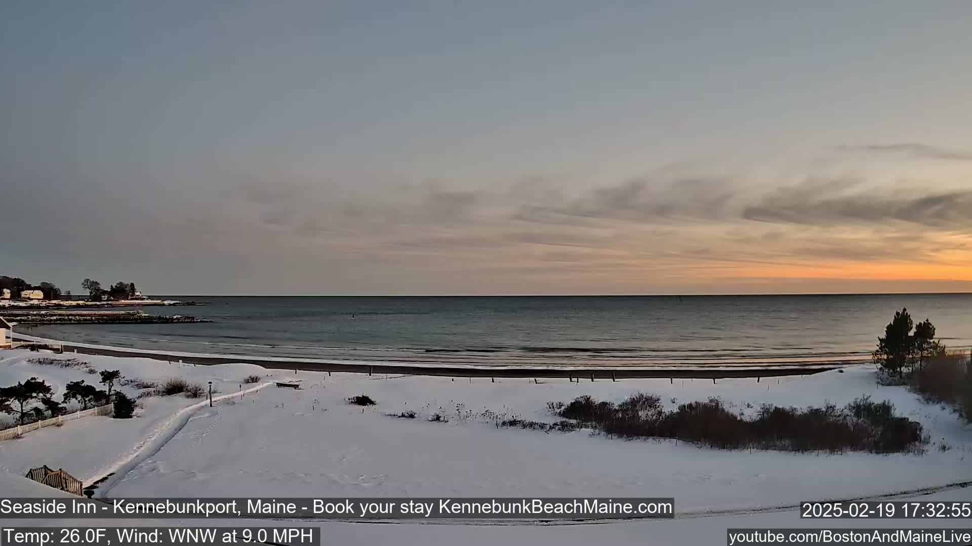 A snow-covered beach and shoreline at sunset, with a calm ocean and a light wind.