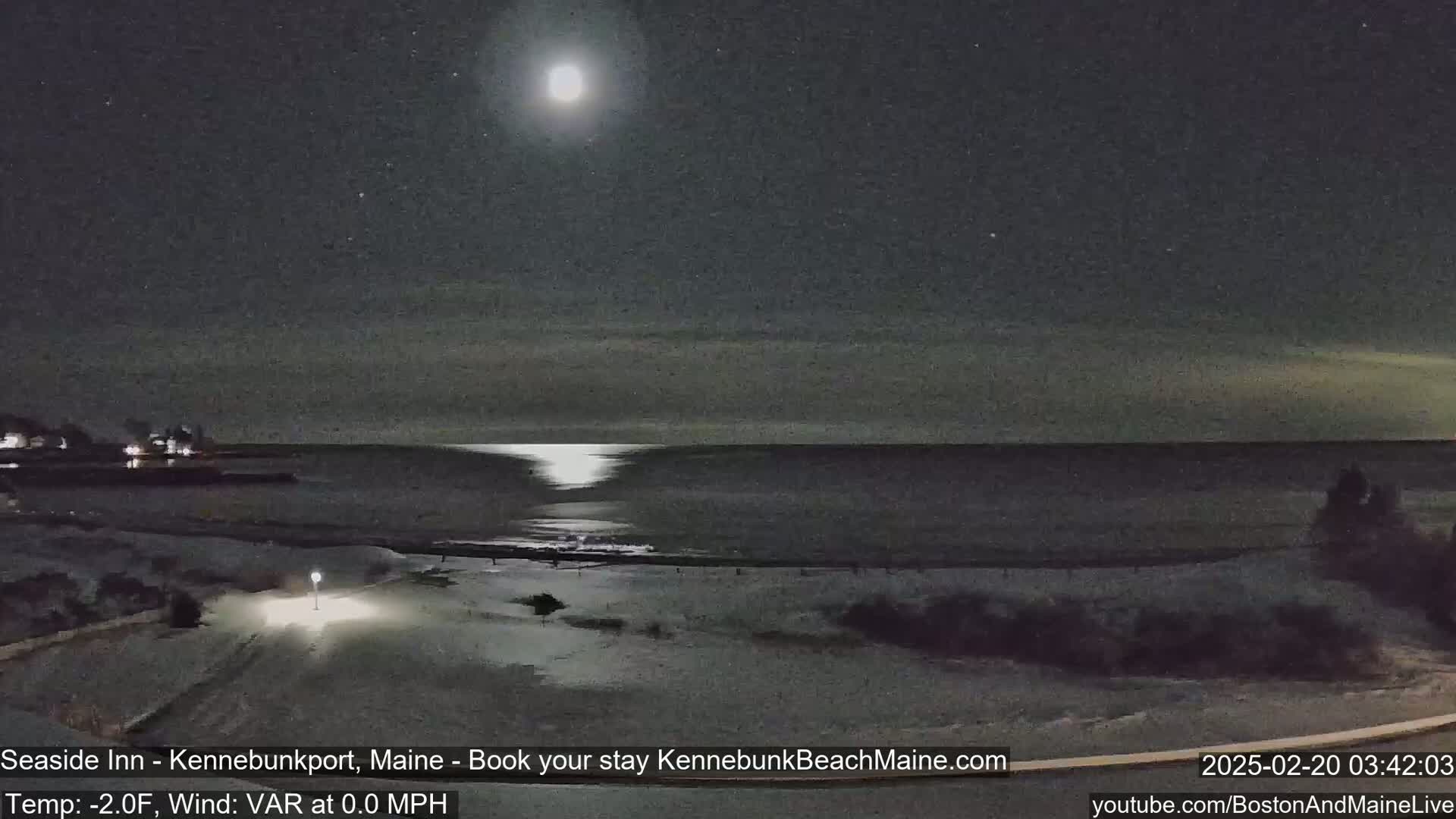 A bright moon illuminates a snow-covered beach and calm ocean at night.
