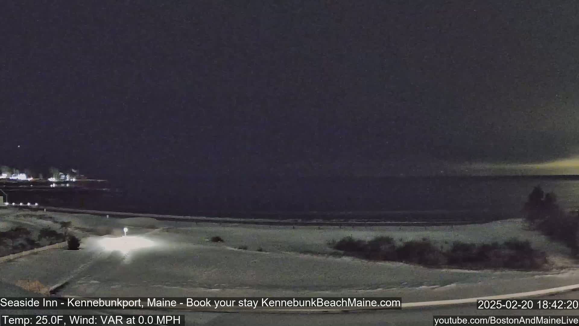 A nighttime, snow-covered coastal scene shows a dark ocean, a beach, and a roadway under a mostly clear, dark sky.
