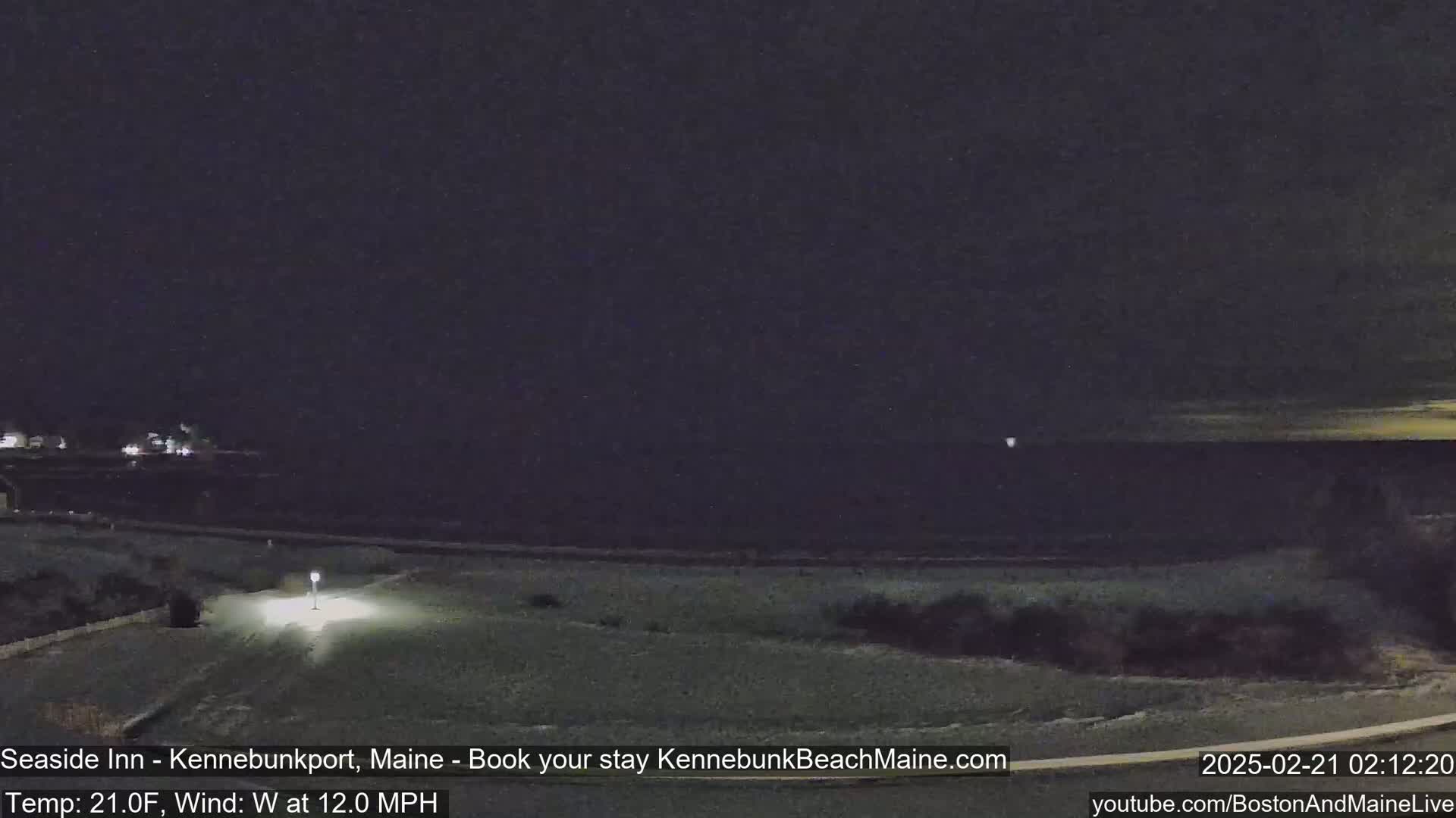 A dark, snowy landscape at night shows a body of water under a mostly clear sky, with a faint light on the horizon and a single light illuminating a small area of land in the foreground.