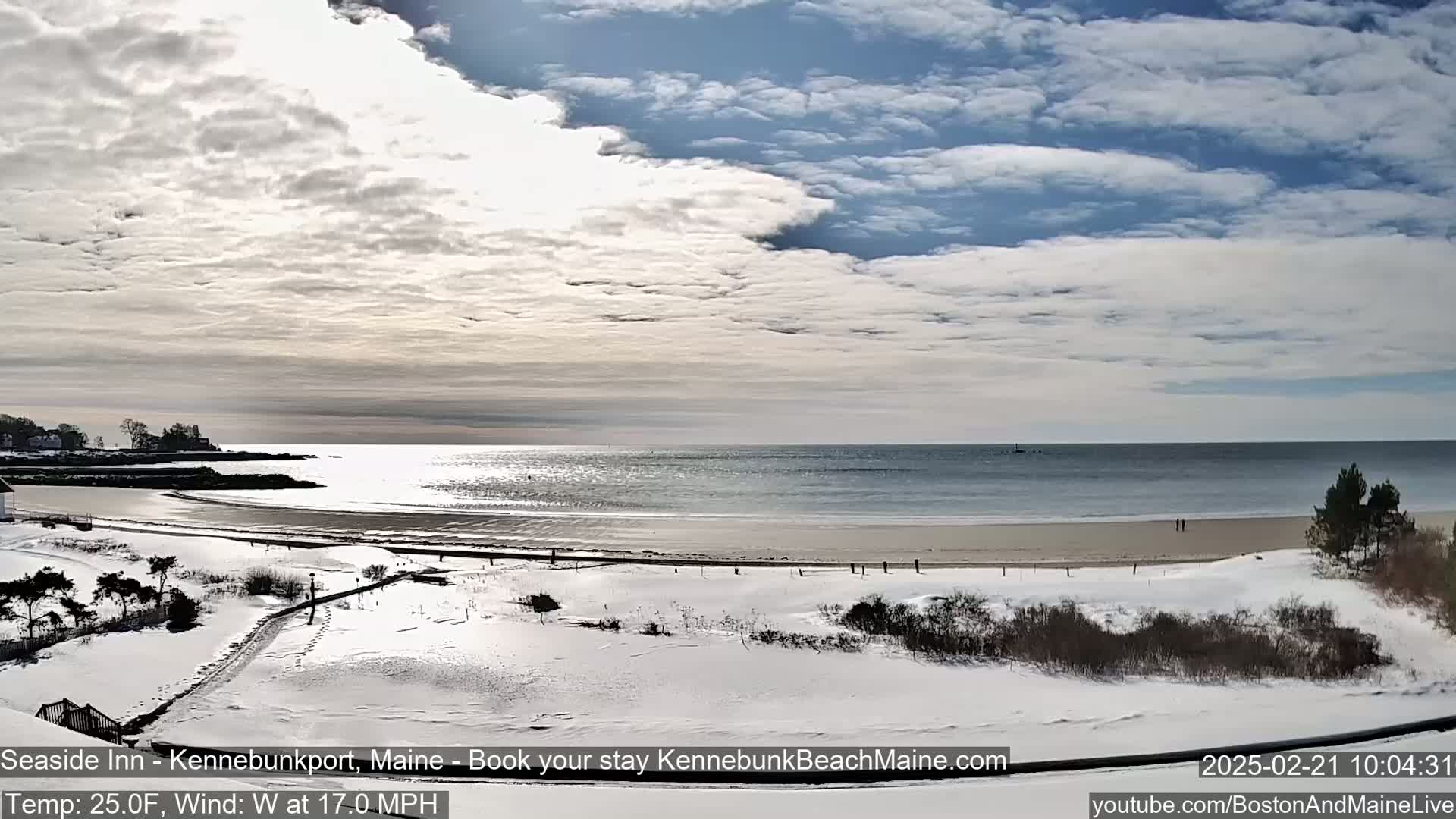 A snow-covered beach stretches along a calm ocean under a partly cloudy sky.