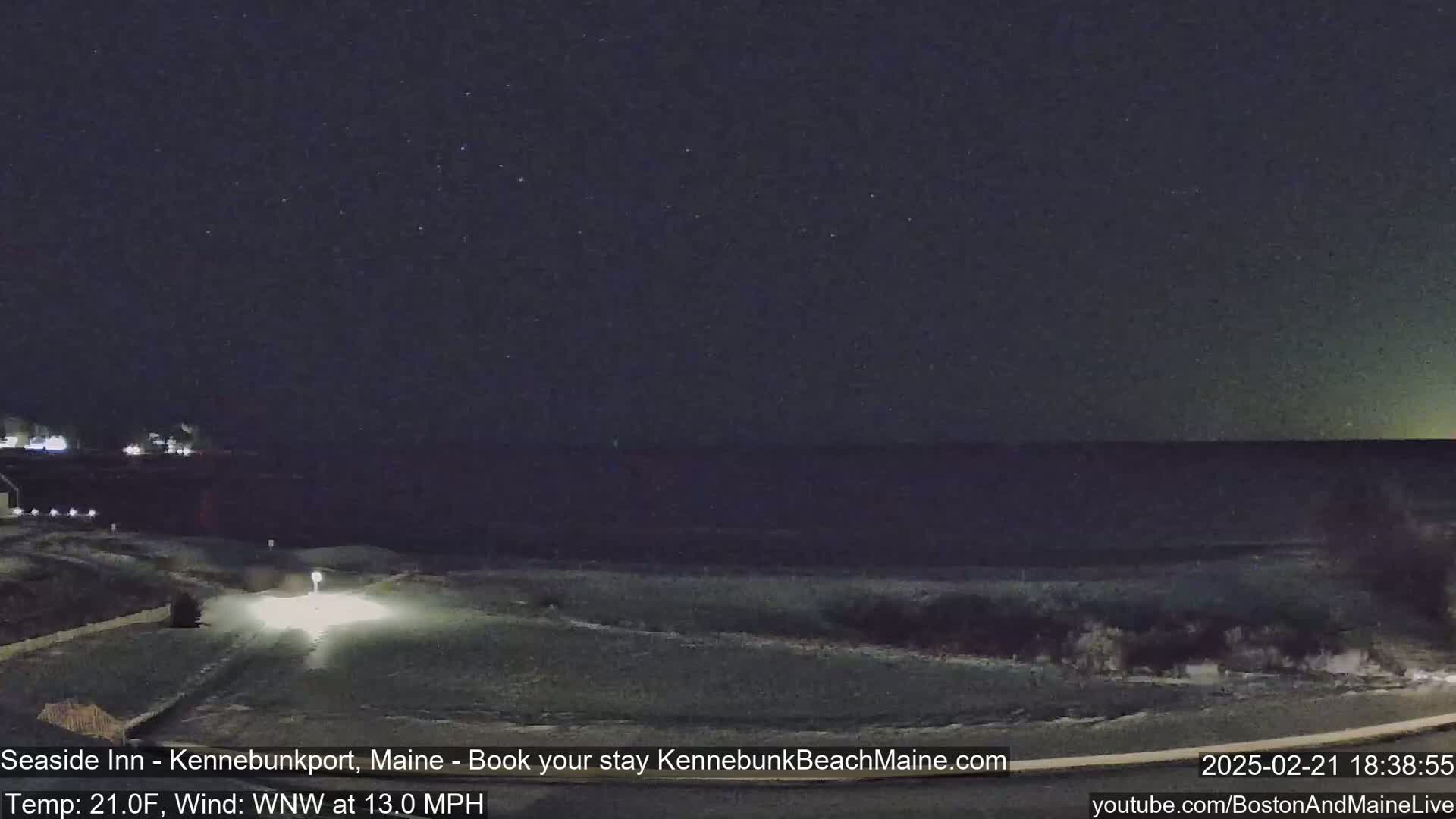 A nighttime view of a snow-covered shoreline with a vehicle's headlights visible on a road leading towards the water under a clear, starry sky, with a wind of 13 mph.