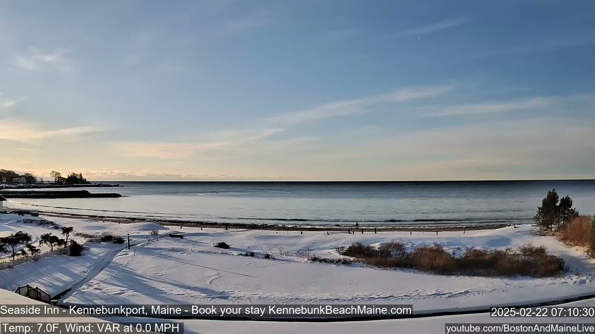 A snow-covered beach and grassy area extend to a calm ocean under a clear, light blue sky.