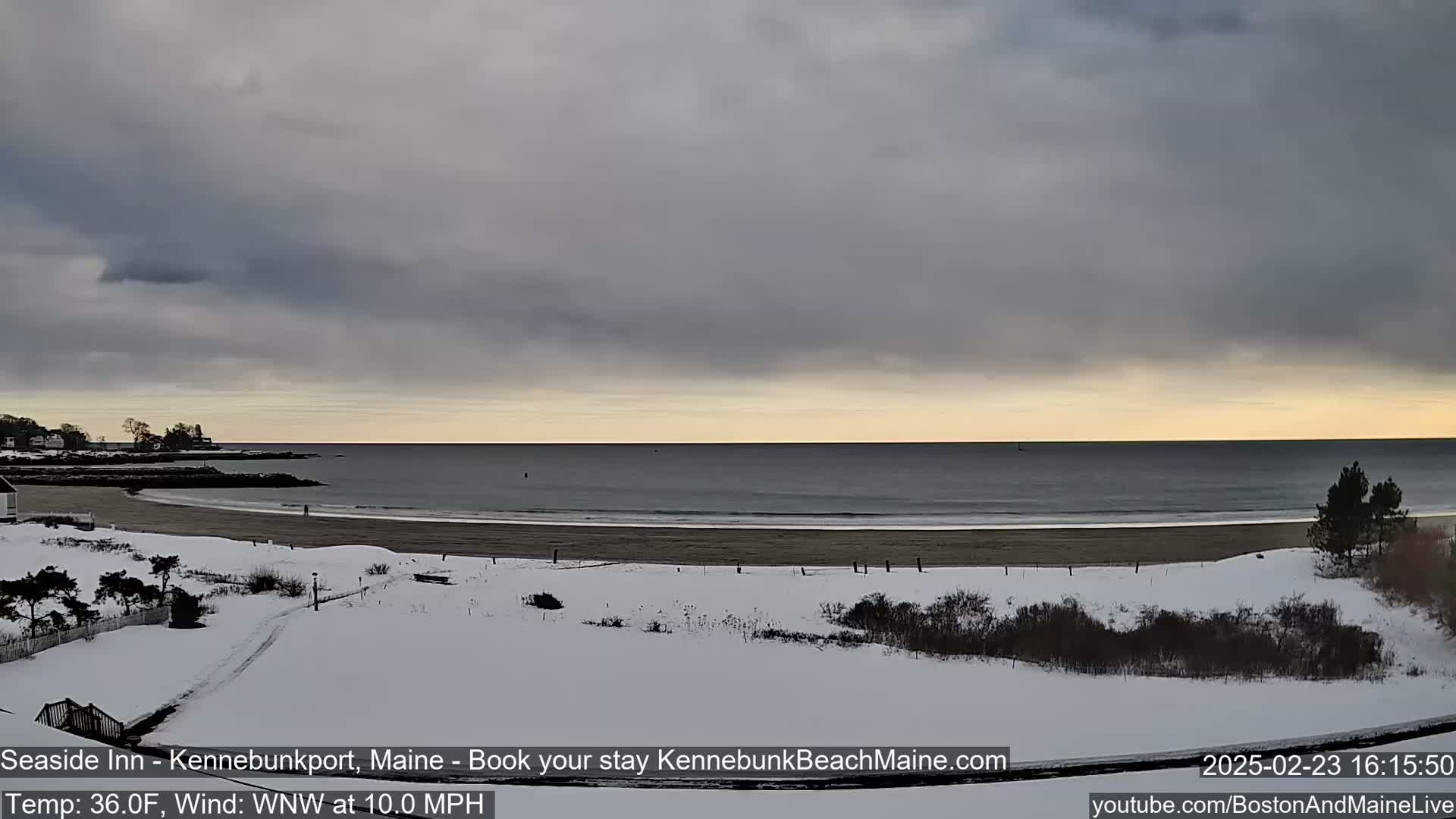 A snow-covered beach and dune landscape stretches to a calm ocean under a mostly cloudy sky.
