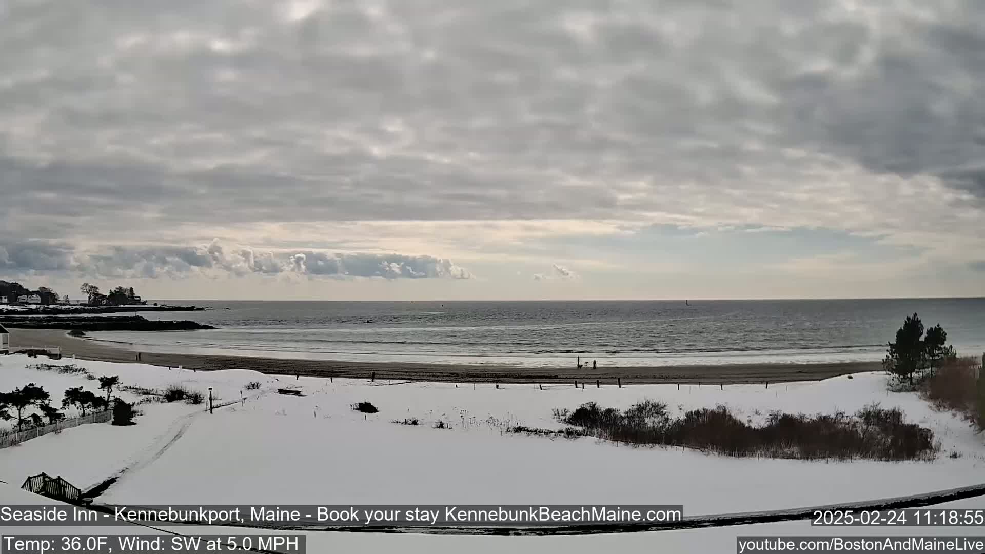A snow-covered beach and grassy area overlook a calm ocean under a mostly cloudy sky.