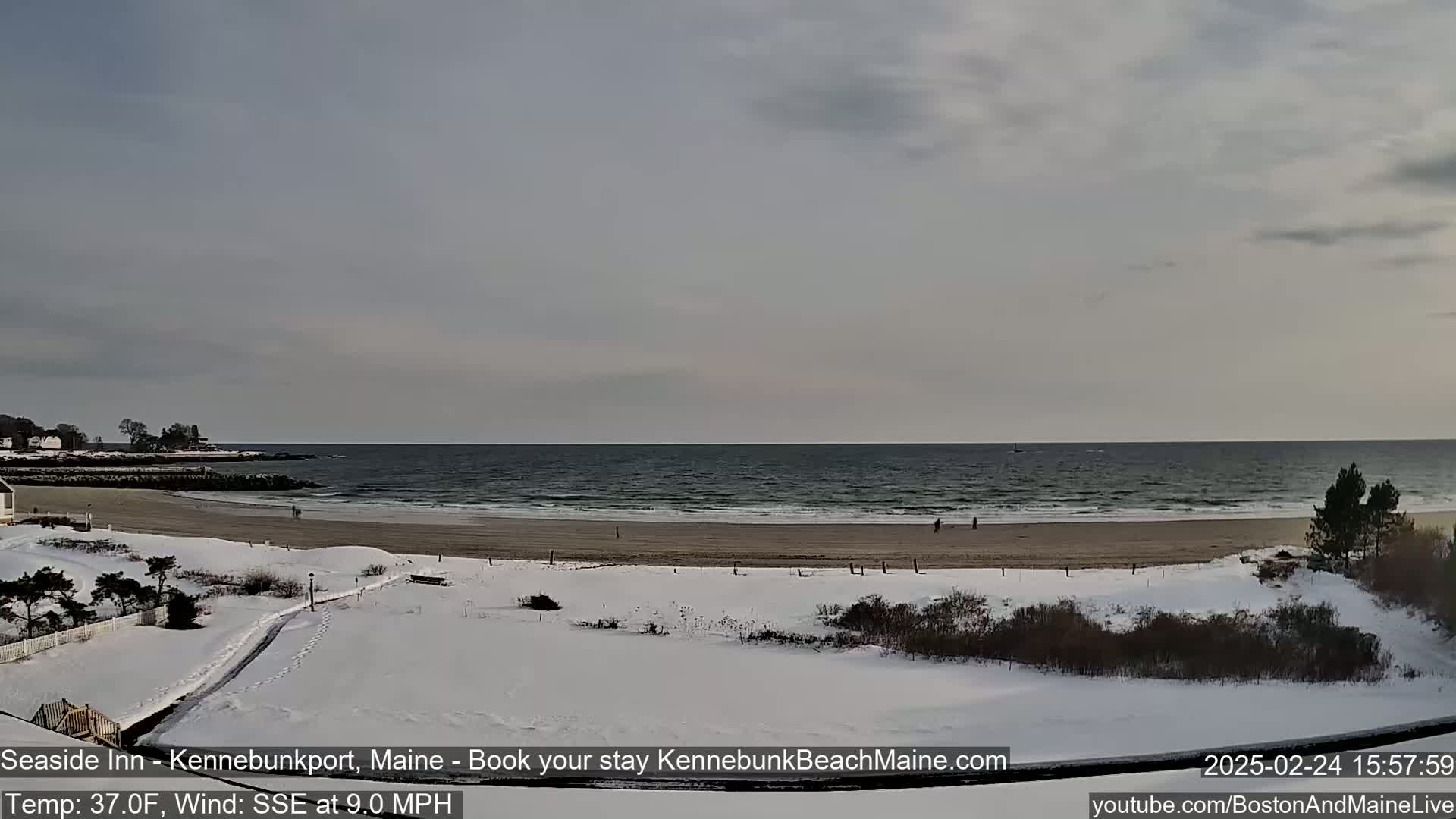 A snow-covered beach with a few people walking along the shore, overlooking a calm ocean under a cloudy sky.