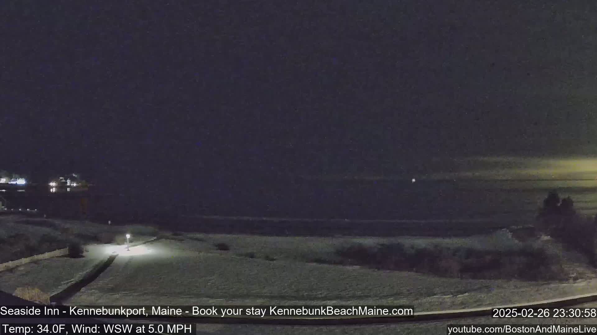 A snow-covered landscape at night shows a dark ocean under a mostly clear sky, with a few lights visible in the distance.