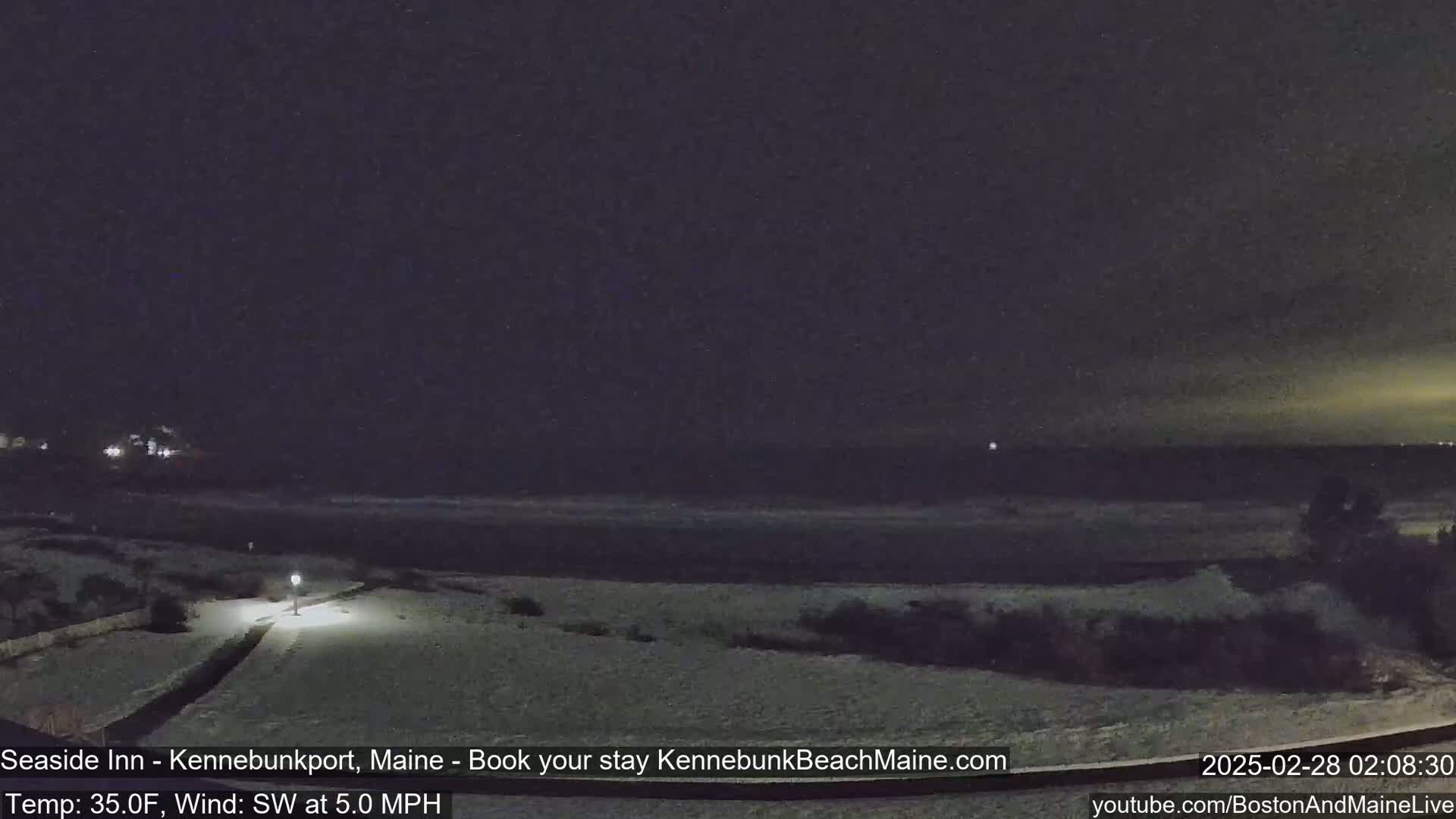 A snow-covered beach at night is seen under a dark sky, with a lamp illuminating a walkway.
