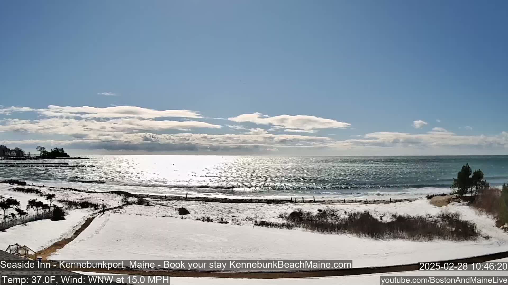 A snow-covered beach with a wavy ocean under a partly cloudy, sunny sky.