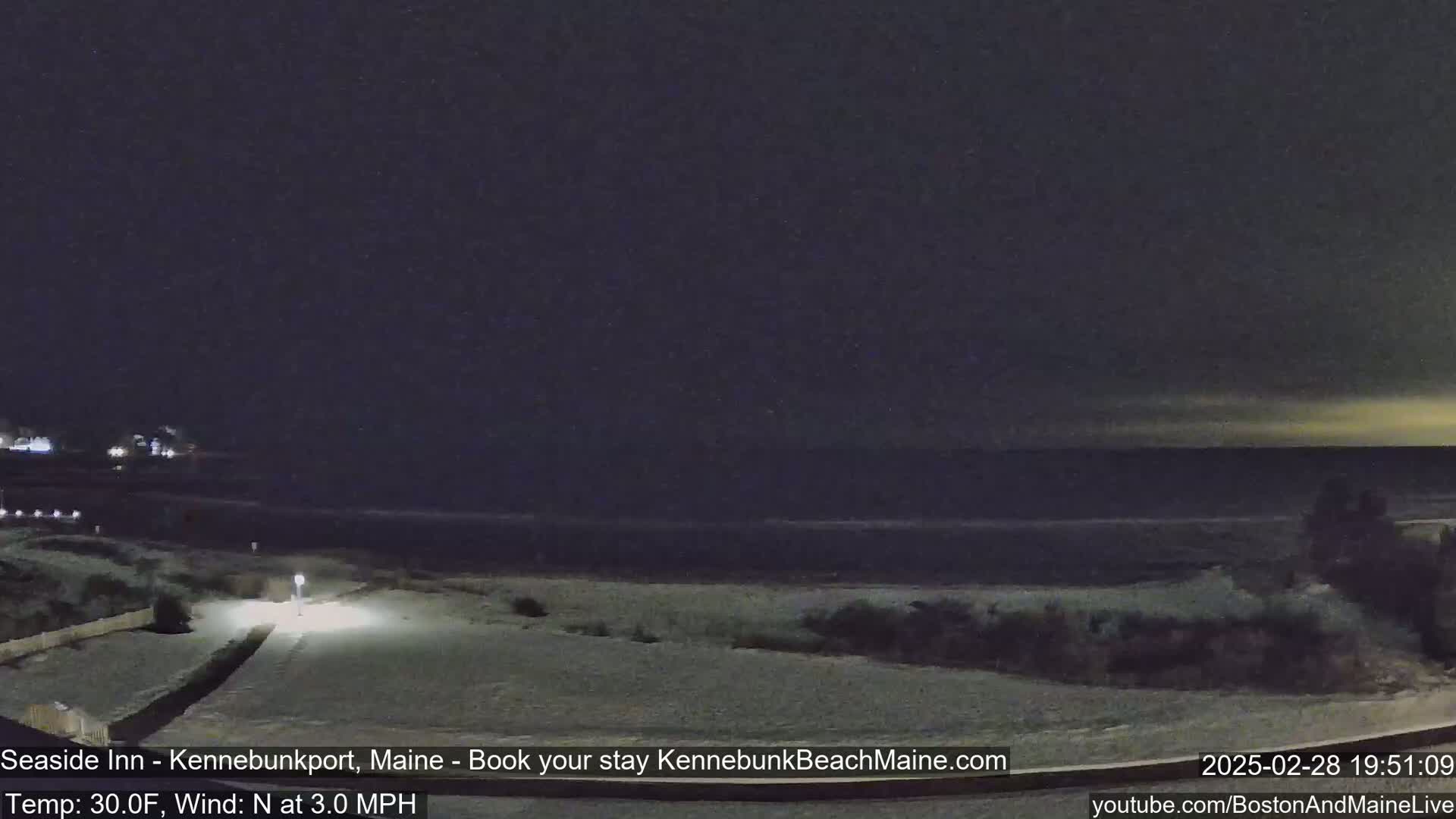 A snow-covered beach at night is seen under a dark sky, with a calm ocean in the background and a lightly lit path leading toward the viewer.