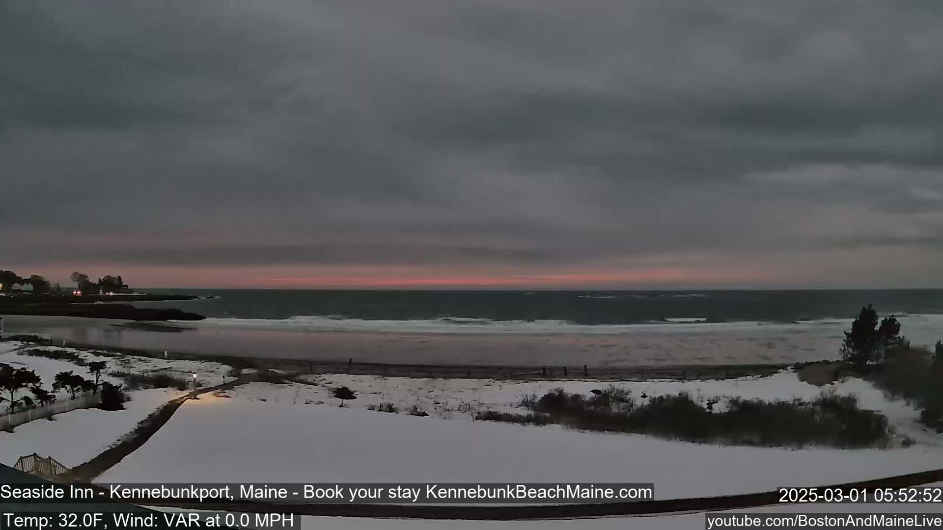 A snow-covered beach scene at dawn shows a calm ocean with a pink and gray sky.