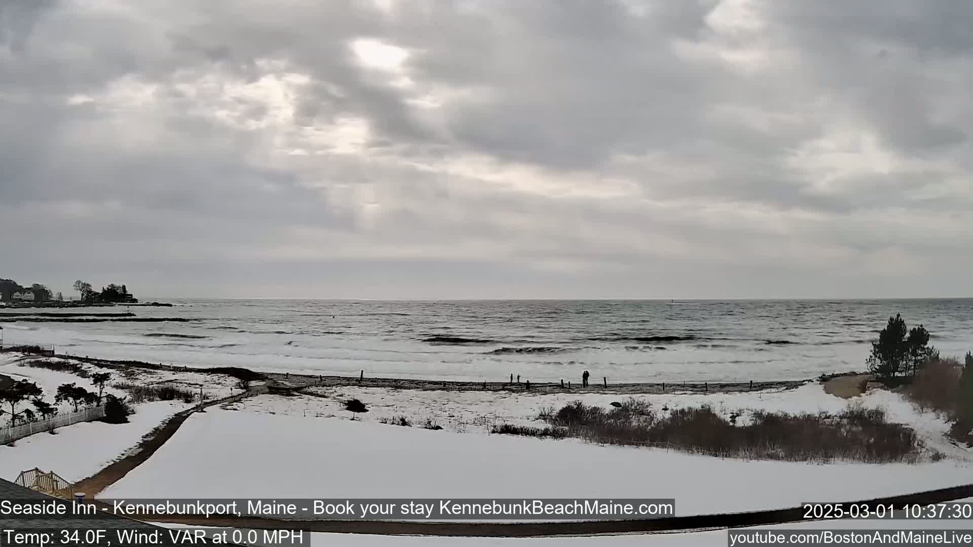A snow-covered beach with a partly cloudy sky and a wavy ocean is visible from a slightly elevated vantage point.