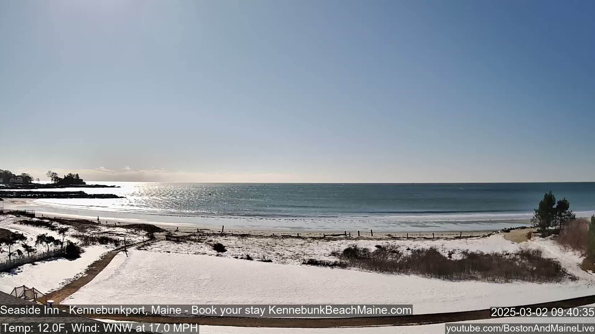 A snow-covered beach with a calm ocean under a clear, sunny sky is visible from a slightly elevated vantage point.