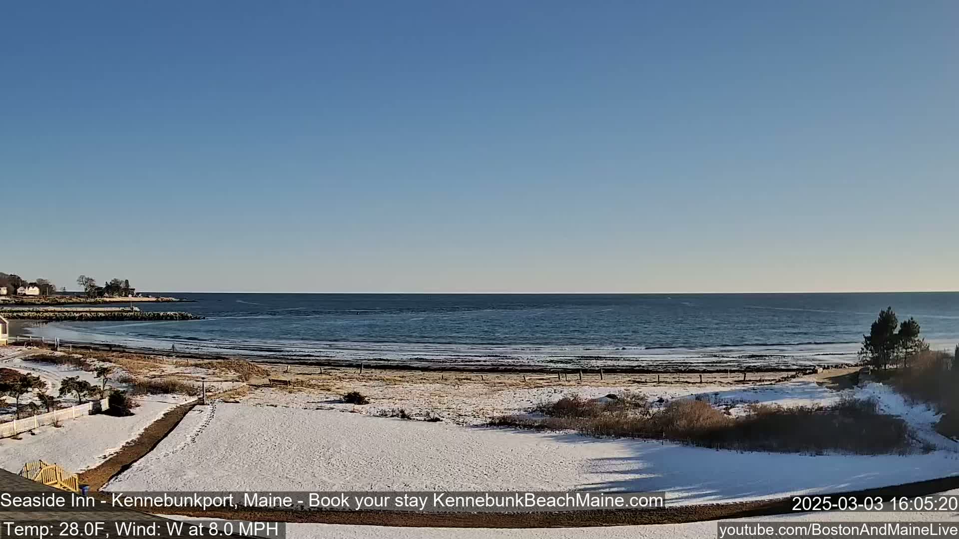 A snow-covered beach with a calm ocean under a clear blue sky is seen from a distance.