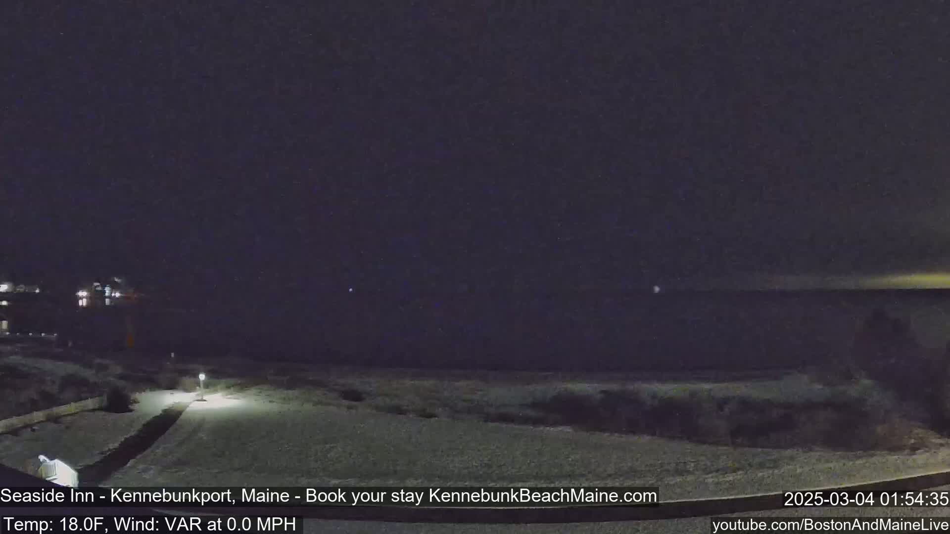 A nighttime snow-covered landscape shows a path leading to a body of dark water under a clear, dark sky.