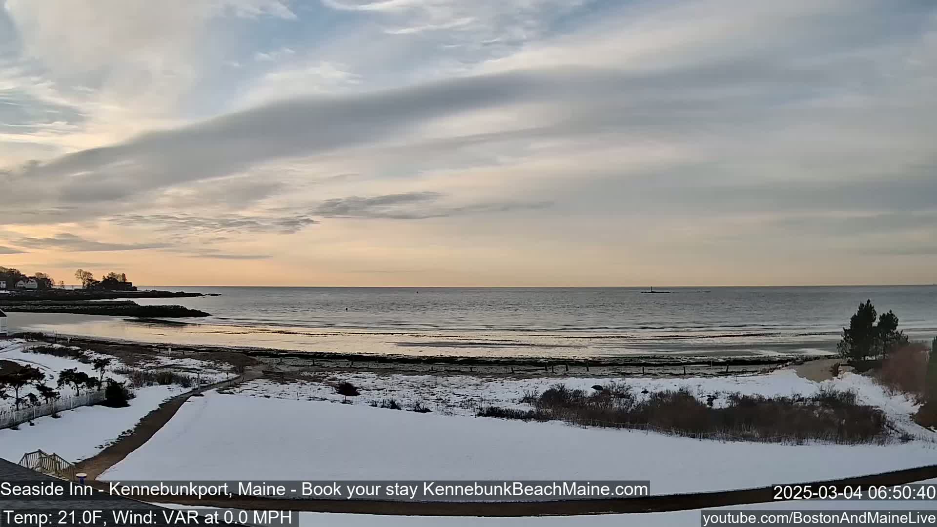 A snow-covered beach and grassy area overlook a calm ocean under a partly cloudy, pale sky at sunrise.