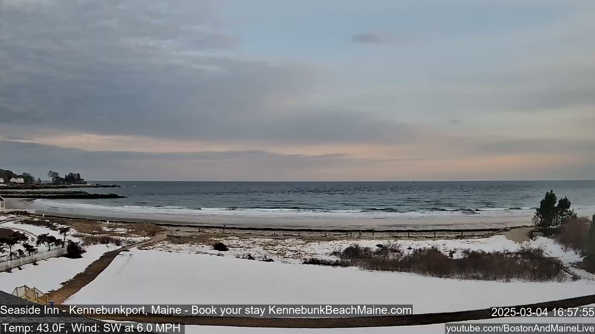 A partly cloudy, cool day shows a wide sandy beach with a calm ocean, partially covered in snow, and vegetation along the shoreline.