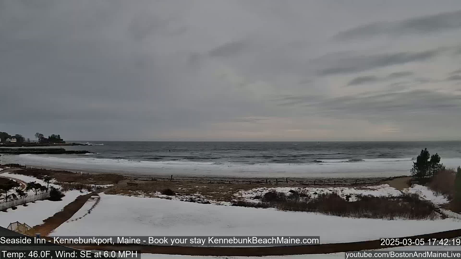 A partially snow-covered beach and coastline are visible under a cloudy sky, with a gray sea showing small waves.
