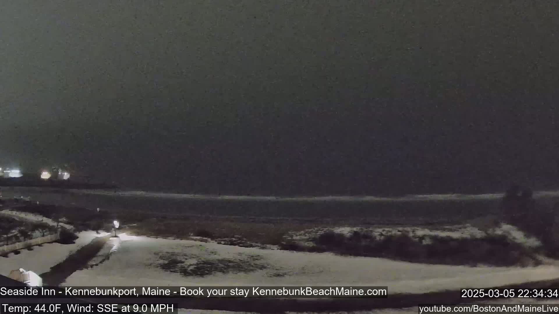 A snow-covered beach at night is shown with a dark, overcast sky and a calm ocean.