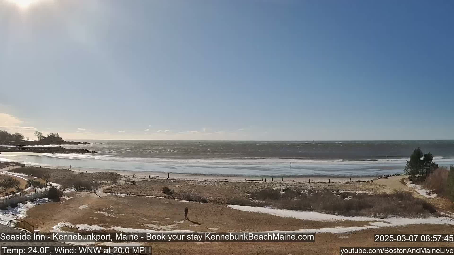 A person walks across a mostly barren, snow-dusted beach overlooking a choppy ocean under a bright, sunny sky.