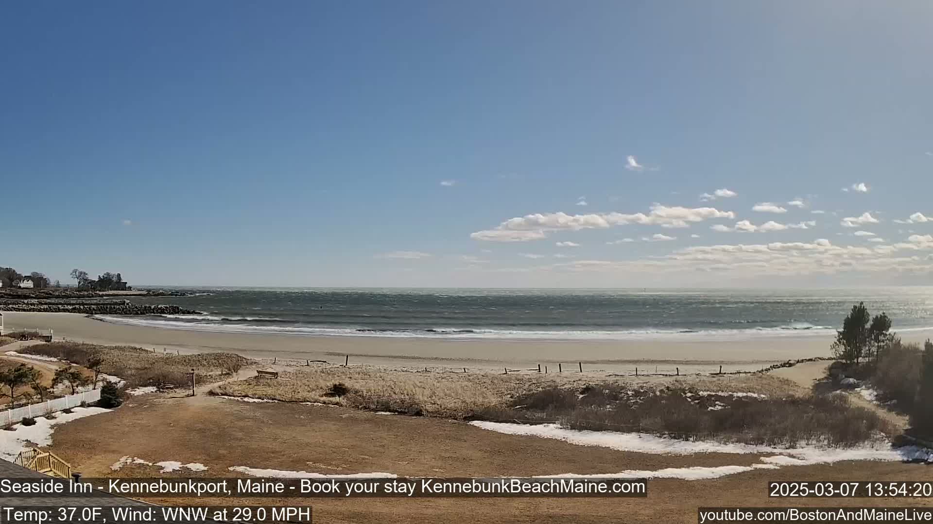 A windy, sunny day at a beach with a sandy shore, crashing waves, and a few patches of snow visible in the foreground.