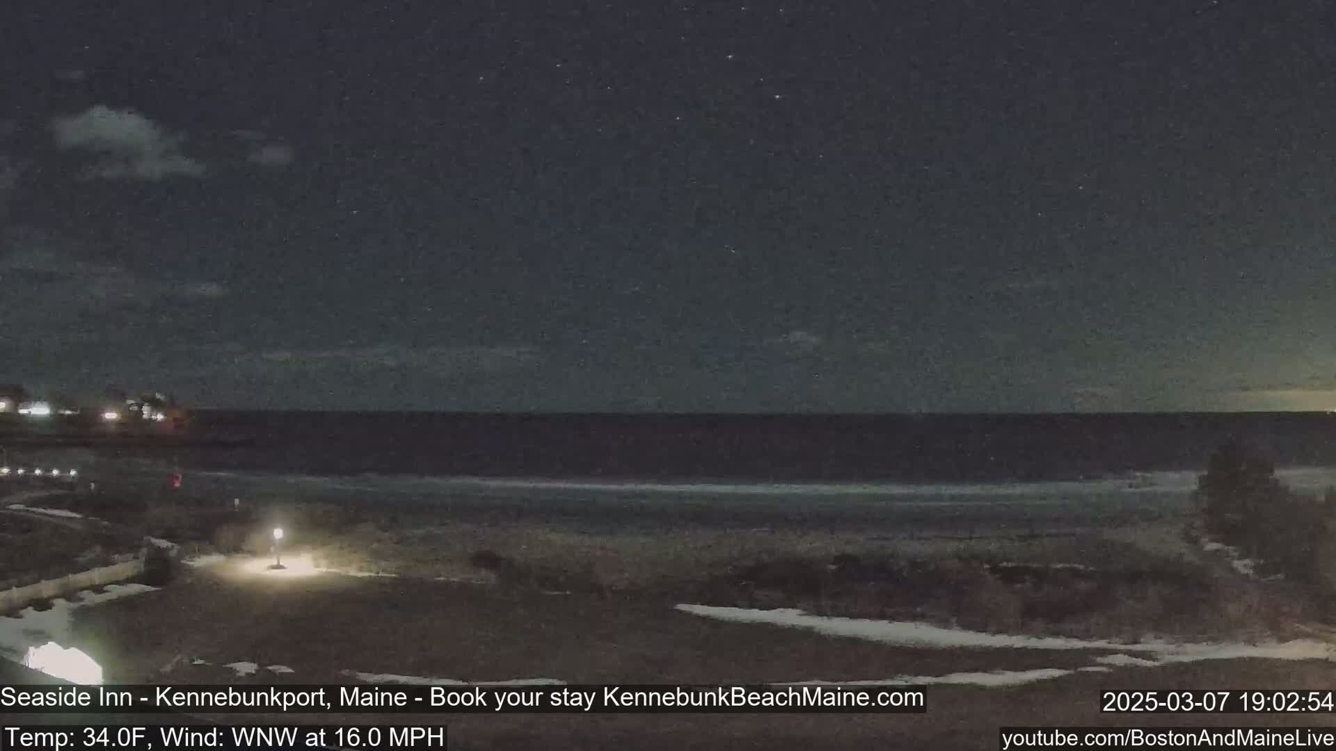 A nighttime view of a dark ocean, a sandy beach with patches of snow, and a paved area with a single light, under a partly cloudy sky.
