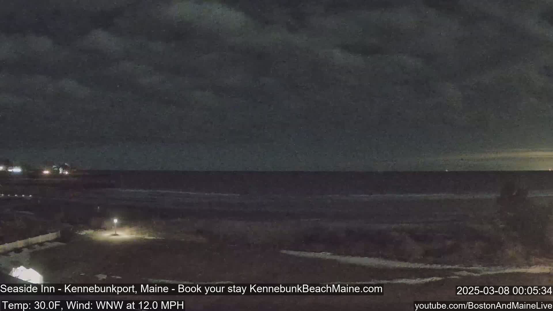 A dark, nighttime beach scene with a mostly cloudy sky and a gently lapping ocean under a wind of 12 mph.