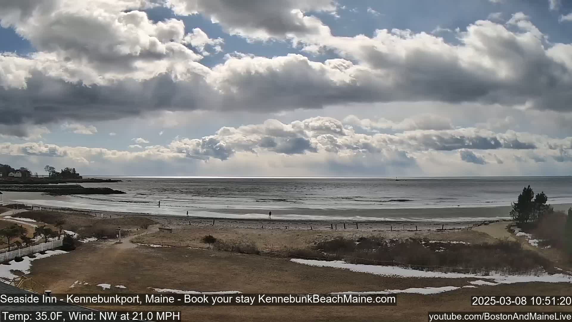 A partly cloudy day with a northwest wind shows a view of a beach with a few people and some snow patches, overlooking a calm ocean.
