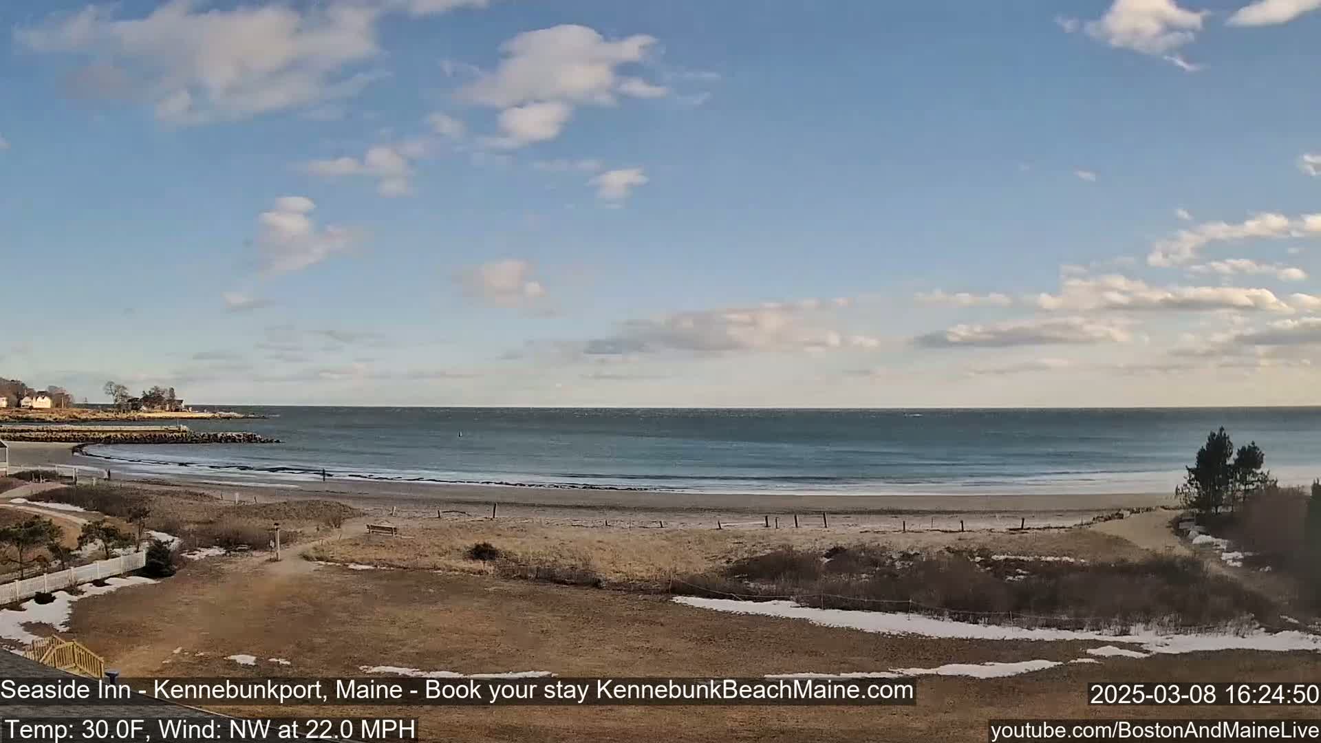 A sandy beach with a calm ocean stretches out under a partly cloudy sky, with a light dusting of snow on the nearby land.
