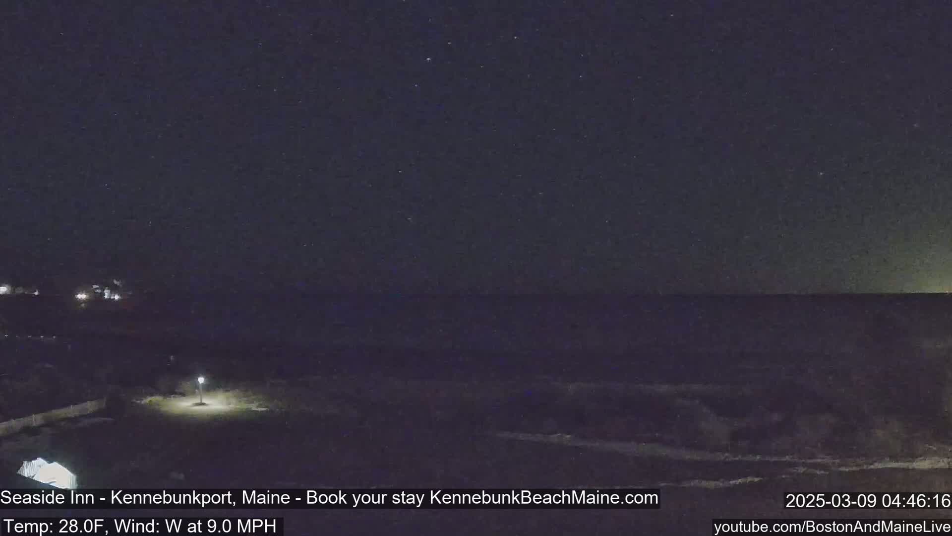 A nighttime view of a dark ocean under a starry sky, with a single lit lamppost visible in the foreground and a slight wind.