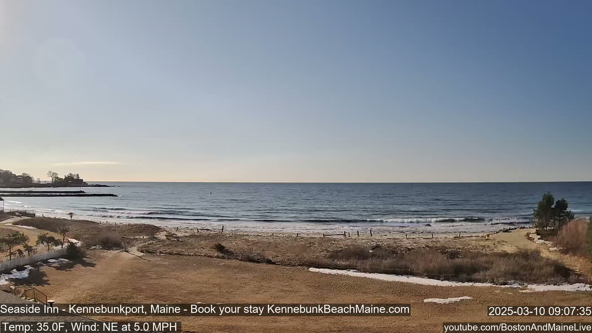 A sandy beach with small waves rolls onto the shore under a clear, bright sky; some patches of snow are visible on the sand and nearby land.