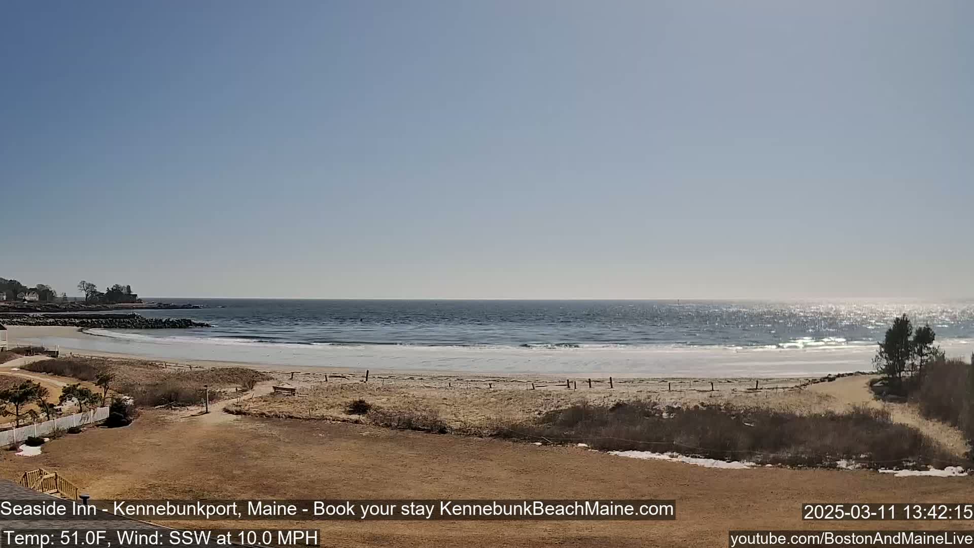 A sandy beach with a calm ocean under a clear blue sky, with some patches of snow visible near the dunes.