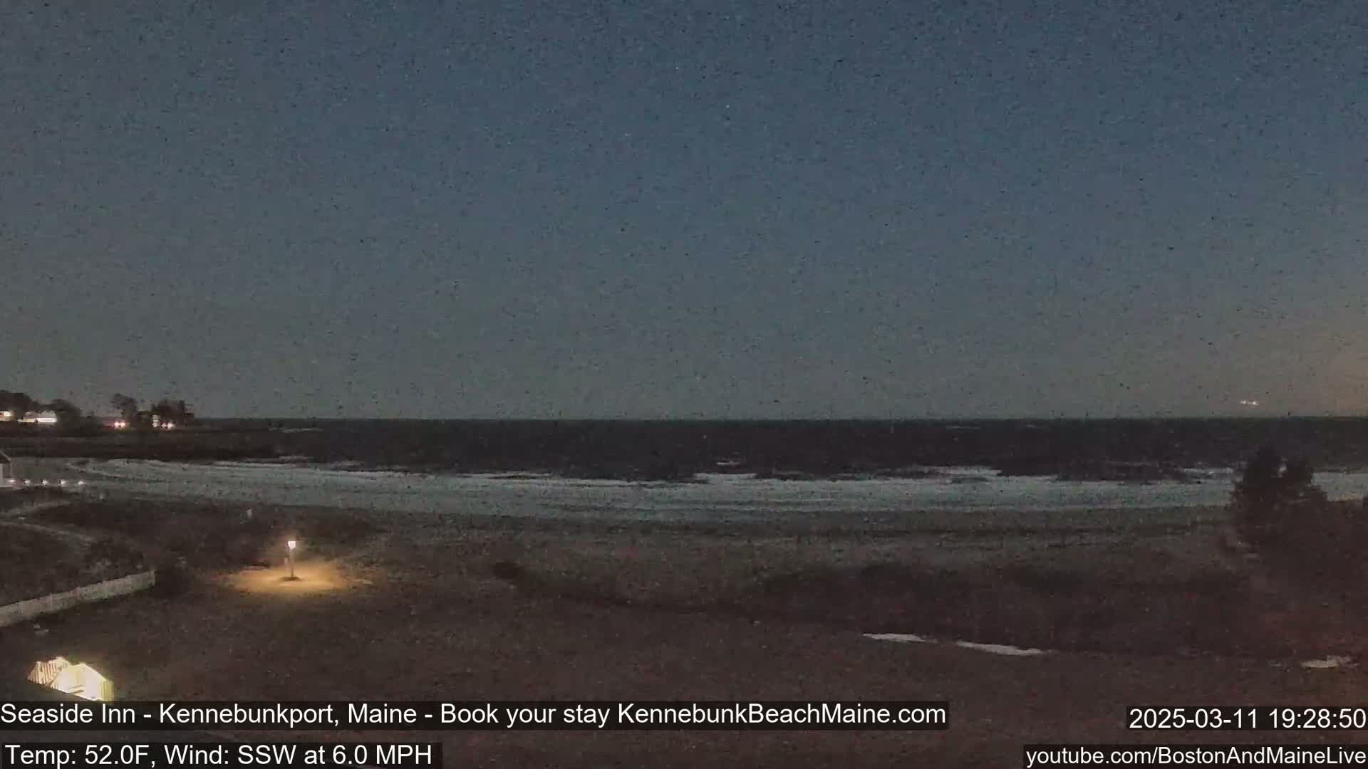 A dark, mostly clear night scene shows a beach with a calm ocean, a single lamppost, and a small amount of snow visible near the shoreline.