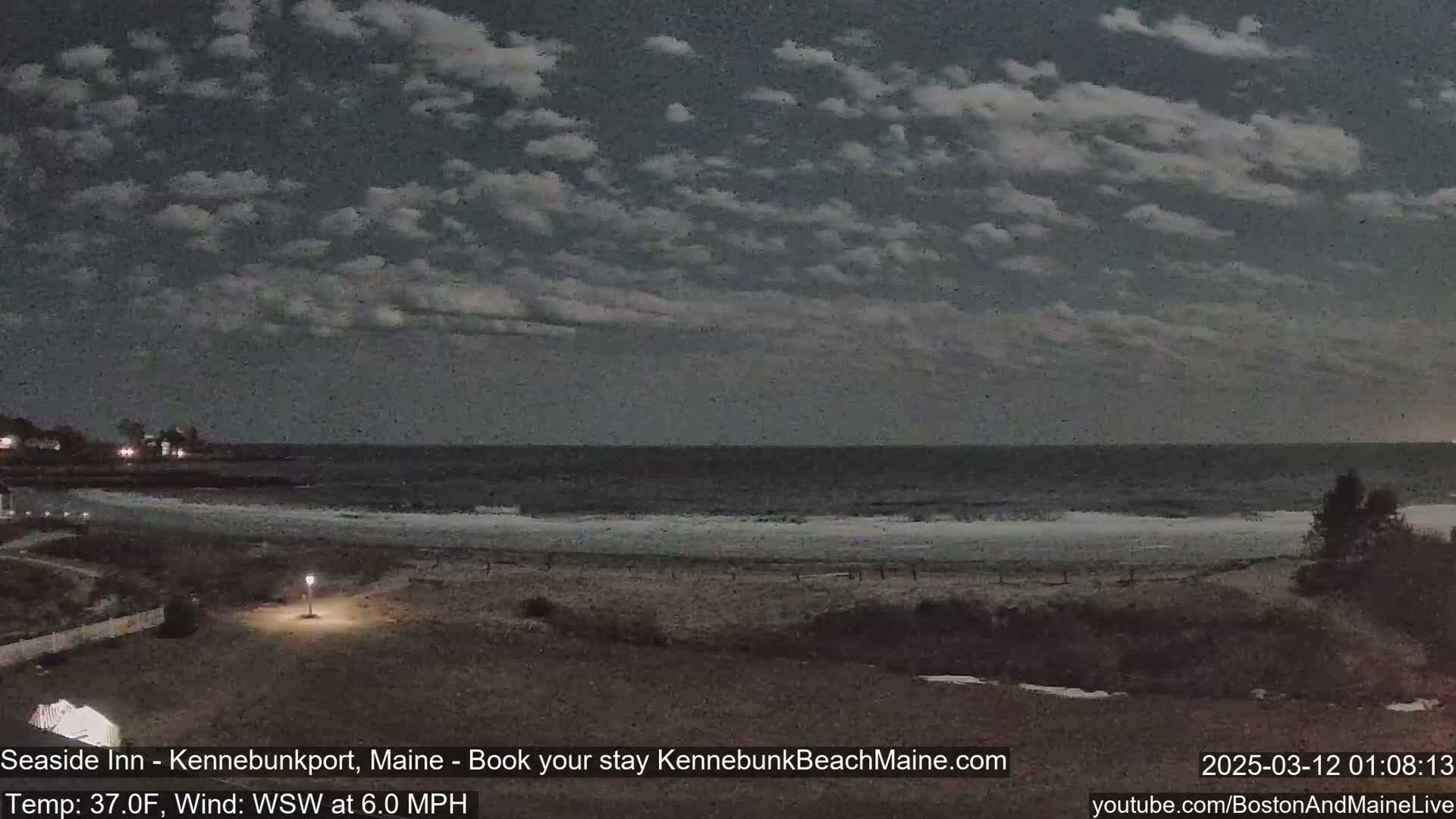 A dark, cloudy night scene shows a beach with a calm ocean under a mostly cloudy sky.