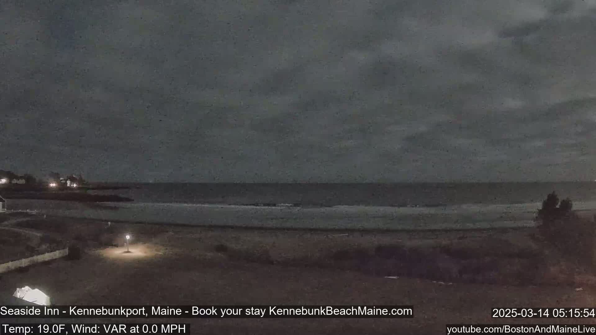 A dark, overcast night scene shows a calm ocean meeting a sandy beach, with a sparsely lit shoreline in the foreground.