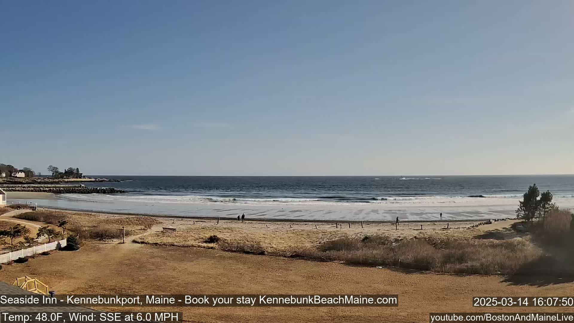A sandy beach with a few people walking stretches out to a calm ocean under a clear blue sky.