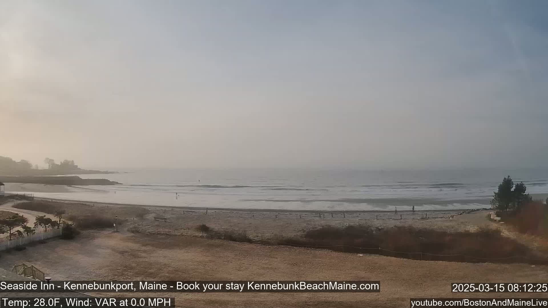 A foggy beach scene shows a calm ocean with small waves lapping a sandy shore.