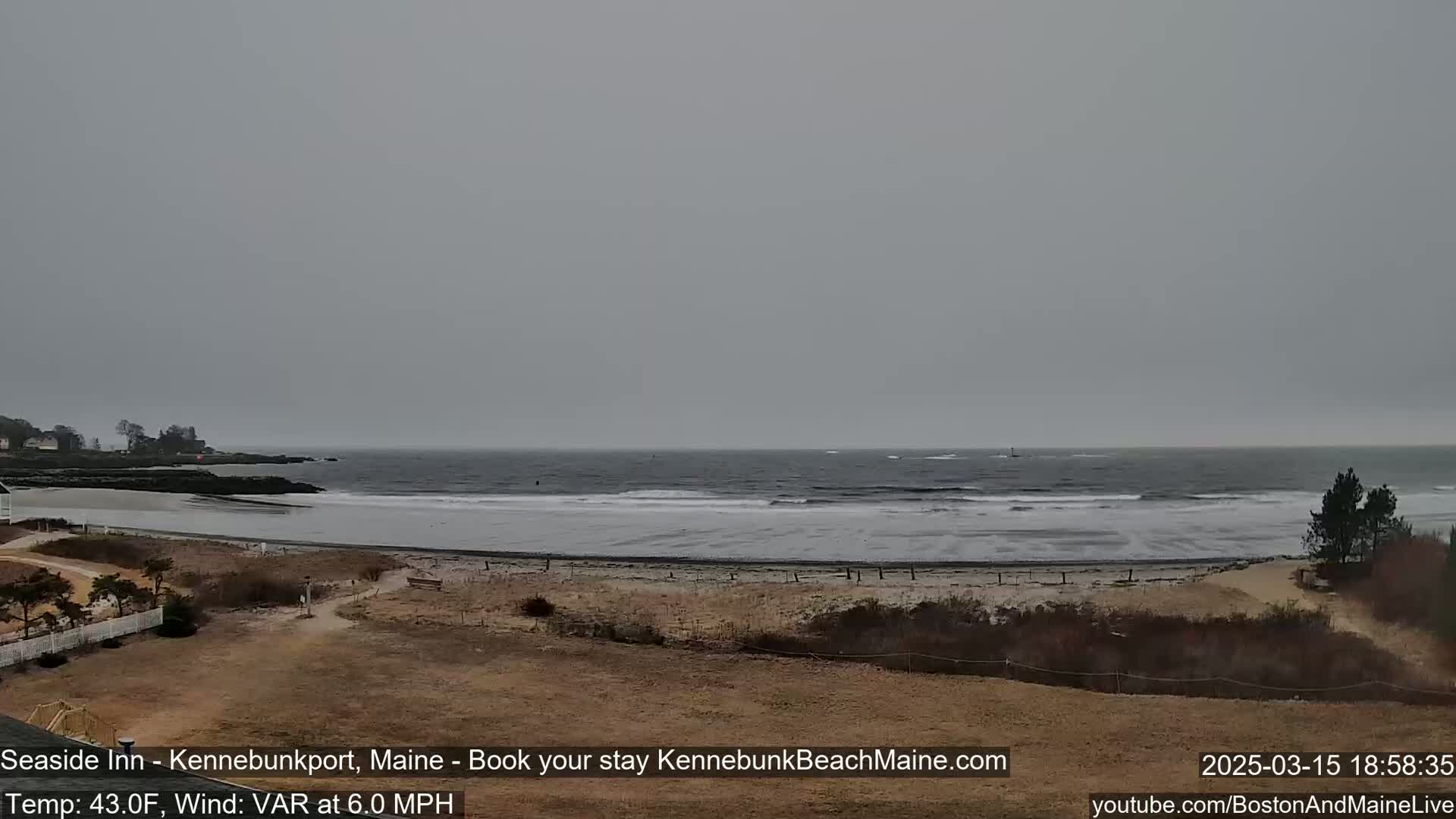 A gray, overcast day shows a beach with small waves lapping the shore, and a flat expanse of sand leading to a rocky point in the distance.