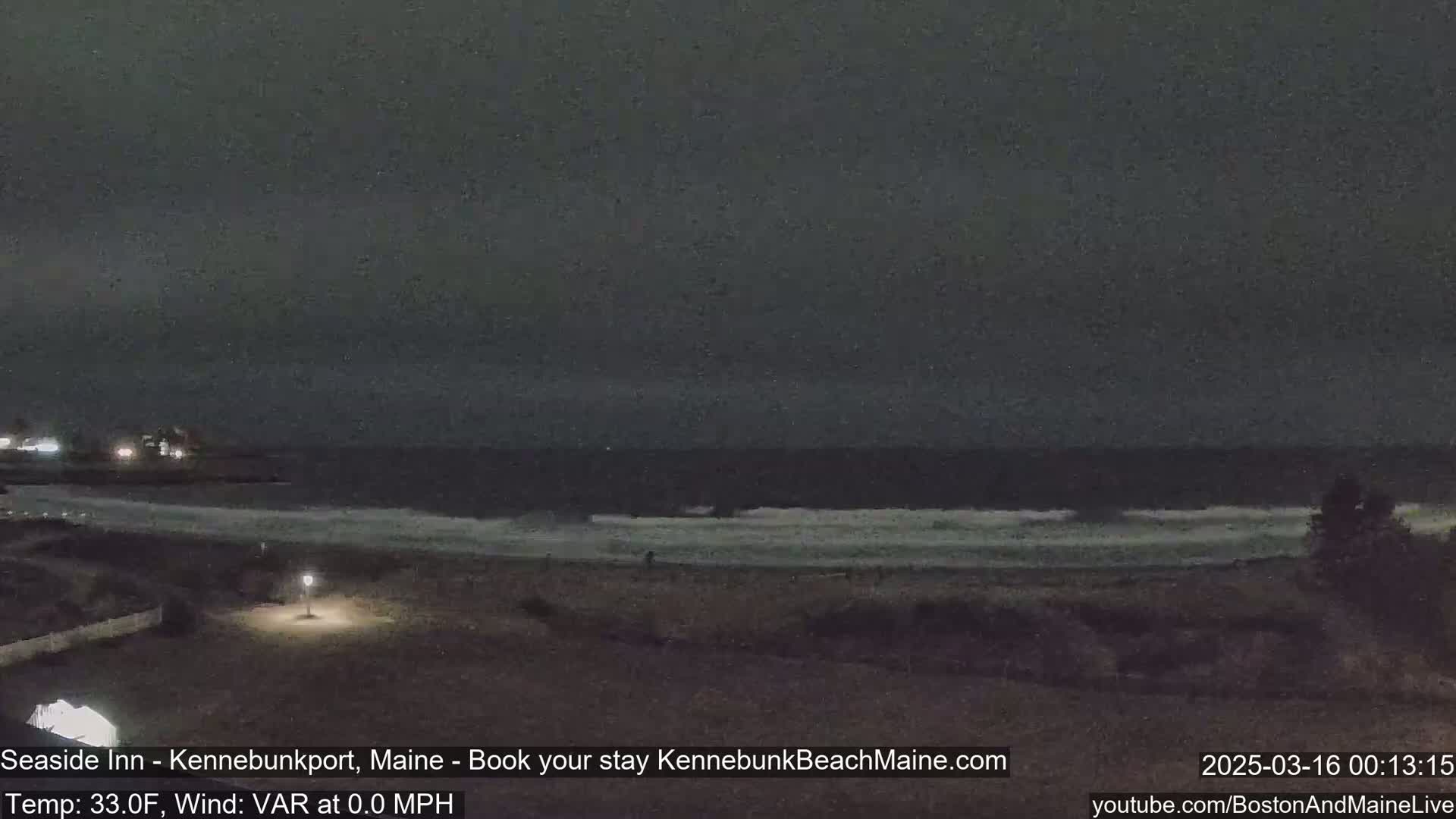 A dark, nighttime beach scene shows a calm ocean with small waves gently lapping the shore under a cloudy sky.