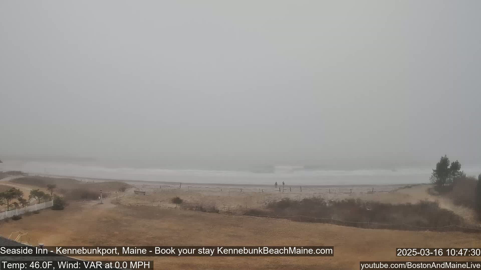 A foggy beach scene shows a stretch of sandy shoreline with small waves and a few people visible in the distance.