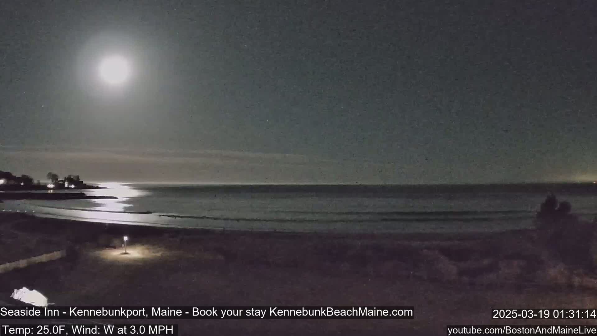 A bright moon illuminates a dark beach and calm ocean at night, with a slight wind.
