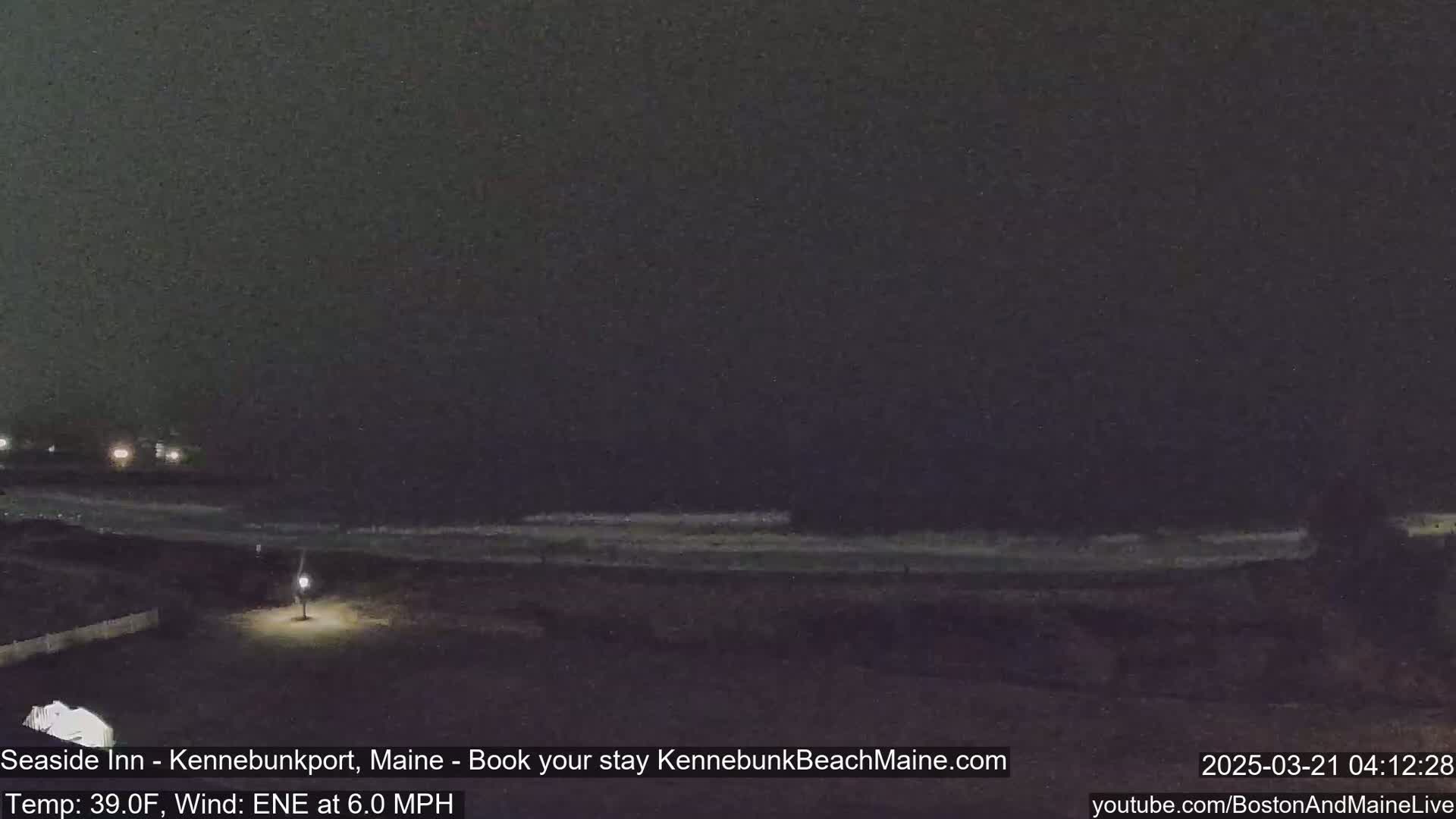 A nighttime view of a dark beach with a calm ocean under a cloudy sky, with a single light illuminating a nearby area.