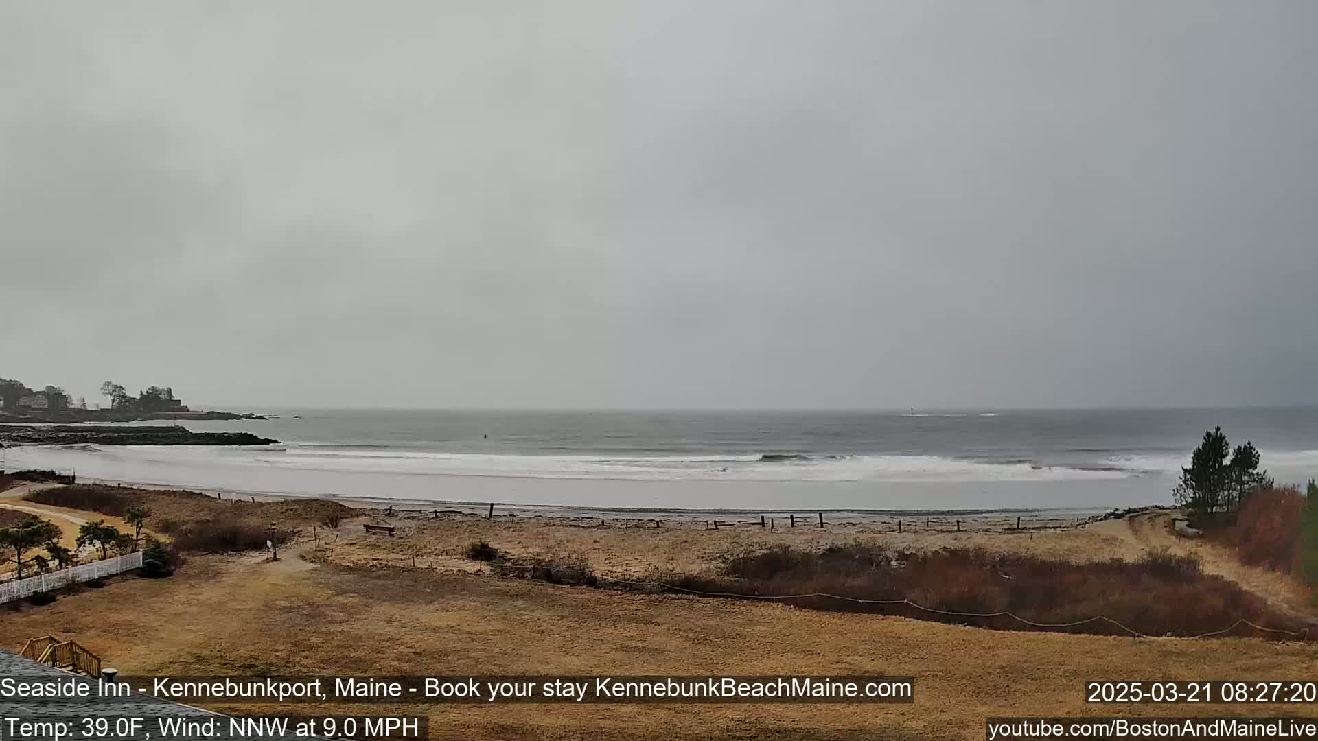 A wide, sandy beach with small waves rolling onto the shore is viewed from a distance on an overcast day with a northwest wind.