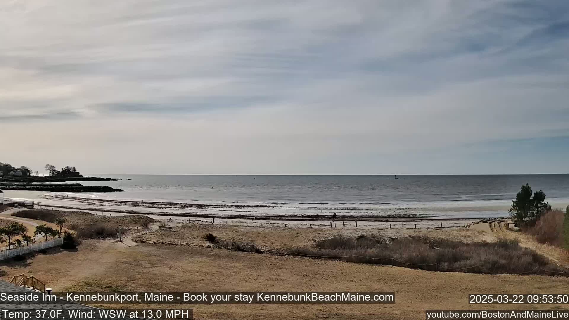 A wide shot of a beach and ocean under a mostly cloudy sky shows a calm sea with a few people walking on the sandy shore.