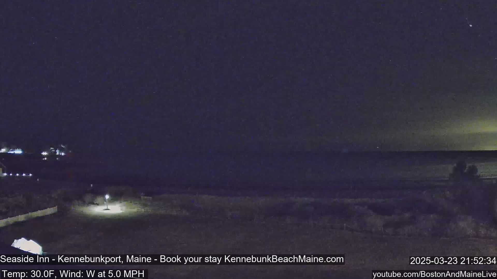 A nighttime view of a dark ocean under a mostly clear, starry sky, with a sparsely lit shoreline in the foreground.